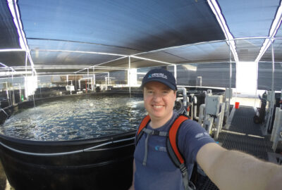 Matt takes a selfie in front of aquaculture operations in Hawaii. A large tank filled with water is behind him.