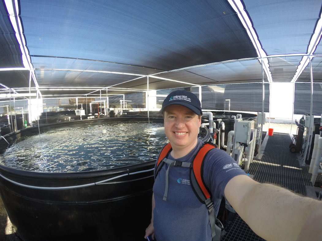 Matt takes a selfie in front of aquaculture operations in Hawaii. A large tank filled with water is behind him.